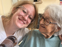 Eine Pflegekraft lächelt neben einer älteren Dame, die in einem Rollstuhl sitzt. Beide sehen freundlich aus und strahlen Freude aus. Die Pflegekraft hält ein Buch in der Hand. Im Hintergrund sind dekorative Elemente zu sehen.
