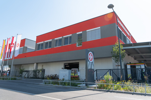 Die Sonne scheint auf den neuen Stützpunkt in der Petritschgasse. Der Stützpunkt hat eine rot-graue, horizontalgestreifte Fassade, bestehend aus jeweils zwei roten und zwei grauen Streifen. Auf der Fassade ist das ASBÖ-Logo sichtbar. Vor dem Stützpunkt stehen drei Flaggen: eine mit dem ASBÖ-Logo, eine Flagge der Stadt Wien und eine österreichische Flagge. Die Szene ist von Sonne beleuchtet, was dem Gebäude einen klaren, hellen Eindruck verleiht