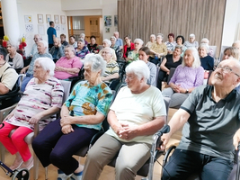 Eine Gruppe älterer Menschen sitzt in einem Raum auf Stühlen. Einige haben Gehhilfen dabei. Die meisten sind aufmerksam und schauen in die gleiche Richtung, während im Hintergrund einige Personen am Stand stehen. Der Raum ist hell und freundlich gestaltet.