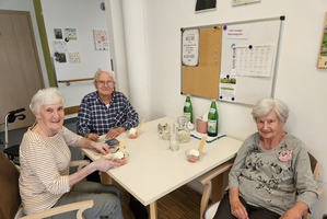 Drei Senioren sitzen an einem Tisch in einem Gemeinschaftsraum. Jeder hat ein Dessert vor sich. Auf dem Tisch stehen auch Glasbehälter und zwei Flaschen Wasser. Die Atmosphäre wirkt gemütlich und gesellig.