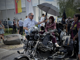 Auf dem Bild steht ein auffälliges motorisiertes Dreirad mit vielen dekorativen Elementen. Eine sitzende Person mit einem Regenschirm wird von mehreren Menschen umgeben, die in einem Freiluftbereich stehen. Im Hintergrund sind Flaggen und parkende Autos sichtbar.