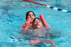 Zwei Jungen im Wasser spielen mit Schwimmnudeln und lachen.