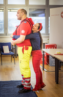 Ein Mann in einer roten und gelben Rettungsdienstuniform steht mit Rücken zu einer Frau, die ihn umarmt. Die Frau trägt eine blaue Bluse mit Punkten und rote Hosen. Im Hintergrund sind Büromöbel und Fenster zu sehen.