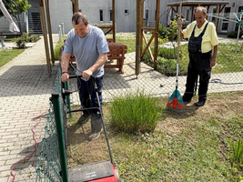 Zwei Männer arbeiten im Garten. Einer benutzt einen Rasenmäher, während der andere mit einem Werkzeug in der Hand wartet. Im Hintergrund sind Gartenmöbel, ein Pavillon und Rasenflächen zu sehen.