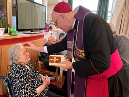 Ein Bischof segnet eine ältere Frau im Rollstuhl. Im Hintergrund sind weitere Personen zu sehen. Der Bischof trägt liturgische Gewänder und hat eine Segenskarte in der Hand. Die Szene vermittelt eine Atmosphäre der Fürsorge und des Glaubens.