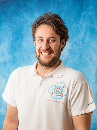 Ein lächelnder Mann in einem weißen Polohemd mit einem Rettungssymbol.