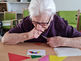 Eine ältere Frau mit Brille sitzt an einem Tisch und betrachtet aufmerksam farbige geometrische Formen. Vor ihr liegen rote, grüne, gelbe, lila und blaue Dreiecke und eine Karte mit einem bunten Muster. Ihr Gesichtsausdruck zeigt Konzentration und Interesse.