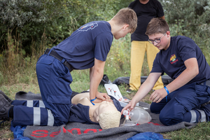 Zwei Jugendliche in Einsatzkleidung üben grundlegende Erste-Hilfe-Maßnahmen an einer Übungspuppe. Der eine hält einen Beatmungsbeutel, während der andere die Puppe stabilisiert. Im Hintergrund sind Bäume und eine weitere Person sichtbar.