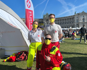 drei Sanaitäter in Uniformen, einer mit roter Jacke, die anderen beiden dahinter mit weißen Polos, auf Wiese vor Hofburg, daneben Einsatzzelt und Beachflag