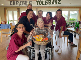 Gruppe von Pflegekräften umarmt eine ältere Frau im Rollstuhl mit Geschenkkorb.