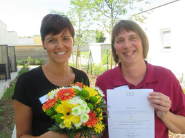 Zwei Frauen halten einen Blumenstrauß und ein Zertifikat in der Hand.