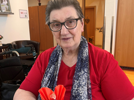 Ältere Frau mit Brille sitzt an einem Tisch und hält eine rote origamiartige Blume mit einer Süßigkeit in der Mitte. Sie trägt ein rotes Oberteil und einen gemusterten Schal. Im Hintergrund sind Stühle und ein Zimmer sichtbar.