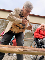 Eine ältere Frau steht über einem Holzstück und bearbeitet es mit einem Werkzeug. Im Hintergrund sitzen weitere Senioren in Rollstühlen, einige blicken auf die Frau. Die Umgebung scheint eine Außenanlage eines Pflegeheims zu sein.