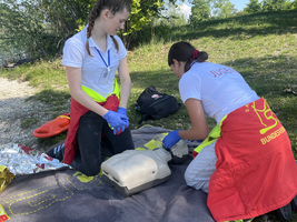 Zwei Jugendliche üben Erste-Hilfe-Maßnahmen auf einer Wiese. Eine Person führt Herz-Lungen-Wiederbelebung an einer Übungspuppe durch, während die andere assistiert. Beide tragen Arbeitshandschuhe und entsprechende Rettungsausrüstung. Im Hintergrund sind Natur und Ausrüstung sichtbar.
