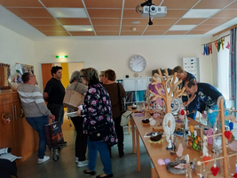 Eine Gruppe von Menschen in einem Raum, der mit Tischen und Stühlen ausgestattet ist. Auf den Tischen sind dekorative Objekte ausgestellt. Einige Personen unterhalten sich, während andere die Ausstellungen betrachten. Die Atmosphäre wirkt lebhaft und gesellig.