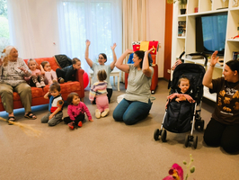 Eine Gruppe von Kleinkindern und Erwachsenen in einem Raum. Die Erwachsenen sitzen oder stehen um einen Sitzbereich, während die Kinder auf dem Boden sitzen und die Hände heben. Ein Kind sitzt in einem Kinderwagen. Es herrscht eine fröhliche und interaktive Atmosphäre.