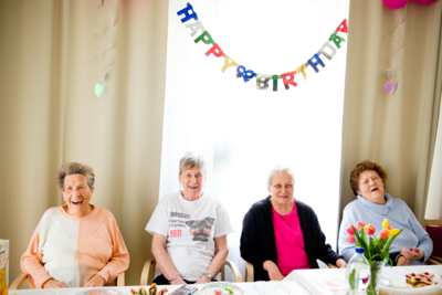 Vier Senioren in Senioren WG sitzen an Tisch und blicken in Kamera, alle vier lachen, im Hintergrund eine Happy Birthday Girlande am Vorhang, auf dem Tisch steht eine Vase mit Blumen