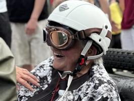 Eine lächelnde ältere Dame trägt einen Motorradhelm und eine Sonnenbrille. Sie sitzt in einem Rennauto und trägt eine gepolsterte Jacke. Im Hintergrund sind Menschen versammelt, die das Geschehen beobachten.