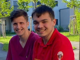 Zwei junge Männer in Arbeitskleidung sitzen lächelnd im Freien.
