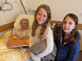 Drei Personen in einem Pflegezimmer: Zwei Mädchen stehen lächelnd neben einer älteren Frau im Bett, die einen Tisch mit persönlichen Gegenständen hat. Die Umgebung wirkt freundlich und hell mit einer Pflanze im Hintergrund.
