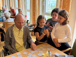 Ein älterer Mann sitzt im Rollstuhl an einem Tisch und spielt mit drei jüngeren Mädchen, die um ihn herumstehen. Auf dem Tisch liegen viele Karten, die für ein Memory-Spiel vorbereitet sind. Im Hintergrund sind andere ältere Menschen zu sehen.