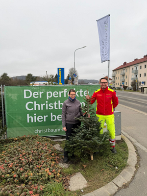 Zwei Personen stehen neben einem Weihnachtsbaum und einem Werbeschild.