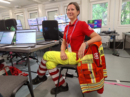 Eine weibliche Person in einem roten T-Shirt und gelben Hosen sitzt in einem Raum mit mehreren Monitoren. Neben ihr liegt eine jacke des Deutschen Roten Kreuzes. Auf den Tischen sind Laptops und Getränke zu sehen.