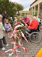 Eine Gruppe von Personen, darunter ältere Menschen und Kinder, versammelt sich um eine mit roten und weißen Bändern geschmückte Stele. Eine Frau im Rollstuhl nimmt aktiv daran teil, während andere zuschauen oder helfen. Im Hintergrund sind Bäume und ein Gebäude sichtbar.