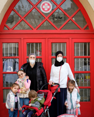 zwei Frauen mit Kopftuch und FFP2-Maske vor roter Tür mit Samariterbund Logo, drei Kinder die bei ihnen stehen, ein Kind in Kinderwagen, blicken alle in Kamera