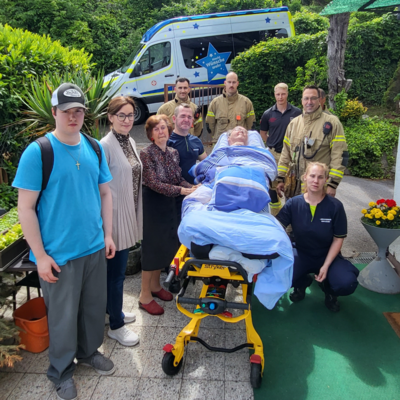 Eine Gruppe von sieben Menschen steht um eine Person, die auf einer Trage liegt, während ein Krankenwagen im Hintergrund zu sehen ist. Einige Menschen tragen Feuerwehr- und Rettungsdienstaffen, während andere zivil gekleidet sind. Die Szene vermittelt eine Atmosphäre der Unterstützung und Besorgnis.
