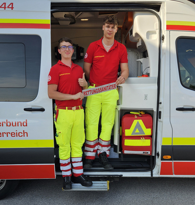 Zwei Rettungssanitäter stehen an einem Rettungswagen und zeigen Daumen hoch.