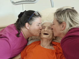 Zwei Frauen küssen eine lächelnde ältere Dame, die im Bett liegt und ein oranges Oberteil trägt. Die Umgebung wirkt freundlich und fürsorglich, was die emotionale Verbindung zwischen den Personen betont.