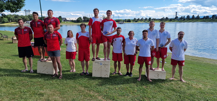 Gruppe von Sportlern in roten und weißen Outfits posiert auf Siegertreppchen am Wasser.