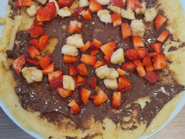Ein flacher Pfannkuchen auf einem weißen Teller, bestrichen mit Schokoladenaufstrich und reichlich mit frisch geschnittenen Erdbeeren und Bananen belegt. Der Pfannkuchen hat einen goldenen Rand und ist appetitlich angerichtet.