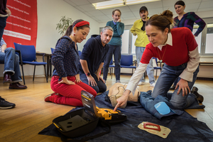 Eine Gruppe von Menschen in einem Raum lernt erste Hilfe. Eine Frau zeigt auf einen Dummy, während andere aufmerksam zuschauen. Es gibt medizinische Ausrüstung wie einen Defibrillator auf dem Boden. Auf der Wand im Hintergrund ist ein Banner mit Informationen zu Erste-Hilfe-Kursen zu sehen.