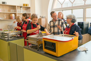 Mehrere Personen stehen in einer Küche und unterhalten sich miteinander. Ein paar von ihnen kochen.