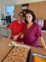 Zwei Frauen stehen an einer Küchentheke und lächeln. Eine Frau trägt ein rotes T-Shirt, die andere ein lila Oberteil. Auf der Arbeitsfläche befinden sich zwei Kuchenbleche, einer mit Streuselkuchen und einer möglicherweise mit einem anderen Kuchen.