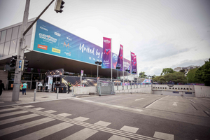 Das Bild zeigt das Eingangstor eines Veranstaltungsortes für den Eurovision Song Contest, der mit bunten Bannern beschmückt ist. Die Umgebung ist verkehrsberuhigt, mit Zebrastreifen und Absperrungen. Im Hintergrund sind Bäume und moderne Gebäude zu sehen.