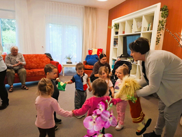 Eine Gruppe von Kindern hält Händchen und tanzt in einem hellen Raum. Ein Erwachsener zeigt ihnen eine Handpuppe. Im Hintergrund sitzen ältere Personen auf einem roten Sofa. Es ist eine fröhliche, interaktive Szene.