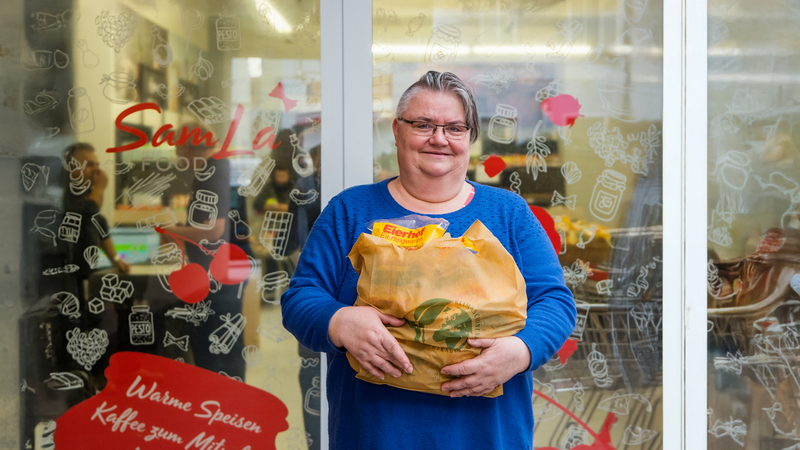 Eine Sozialmarktkundin steht mit einem gefüllten Einkaufssackerl vor dem Sozialmarkt und Lächelt