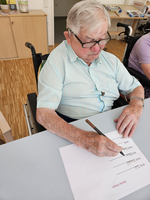 Ein älterer Mann im Rollstuhl schreibt mit einem Stift auf Papier.