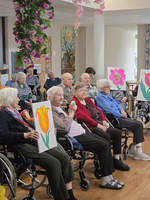 Eine Gruppe älterer Menschen sitzt in Rollstühlen in einem Gemeinschaftsraum. Sie halten Zeichnungen von Blumen in den Händen. Einige Personen lachen und interagieren miteinander, während im Hintergrund dekorative Elemente zu sehen sind.