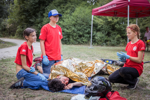 Vier Kinder in roten T-Shirts sind in einer Notfallsituation. Ein Junge liegt auf dem Boden, von einer goldenen Decke bedeckt, während die anderen drei, mit Handschuhen, helfen. Im Hintergrund steht ein Zelt mit einem Logo. Grüne Bäume umgeben die Szene.