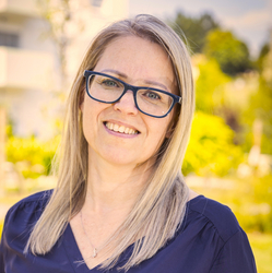 Eine lächelnde Frau mit blonden Haaren und Brille steht im Freien.