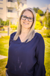 Eine lächelnde Frau mit blonden Haaren und Brille steht im Freien.