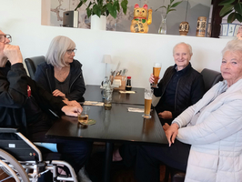 Vier Personen sitzen an einem Tisch in einem Restaurant. Ein Mann im Rollstuhl hält ein Glas, während eine Frau neben ihm sitzt und eine andere Frau in der Mitte lächelt. Der letzte Mann hebt sein Bierglas zum Toast. Im Hintergrund sind Pflanzen und Dekorationen sichtbar.