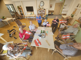 Eine Gruppe älterer Menschen sitzt um einen Tisch in einem Gemeinschaftsraum. Sie halten kleine Fahnen in den Händen und haben lächelnde Gesichter. Im Hintergrund sind Stühle, Pflanzen und ein Fernseher zu sehen. Es herrscht eine freundliche, festliche Atmosphäre.