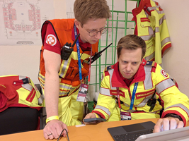 Zwei Rettungskräfte in auffälliger Uniform betrachten gemeinsam einen Laptop, während sie wichtige Informationen austauschen. Im Hintergrund sind Notfallausrüstungen und ein Plan sichtbar. Die Szene vermittelt Teamarbeit und Konzentration in einem Einsatzszenario.