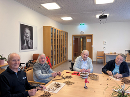 Vier ältere Menschen sitzen an einem Tisch in einem hellen Raum. Sie schälen Nüsse und lächeln. Im Hintergrund hängt ein Porträt einer älteren Person an der Wand. Auf dem Tisch liegen Schalen mit Nüssen und Schalen, die bereits leer sind.