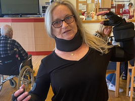 Eine Frau mit Bewegungseinschränkungen zeigt ihre speziellen Handschuhe und Halsstütze.